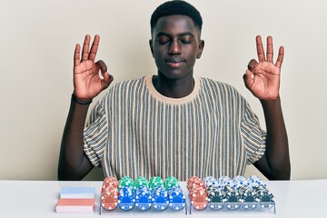 Young african american man sitting on the table with poker chips and cards relax and smiling with eyes closed doing meditation gesture with fingers. yoga concept.