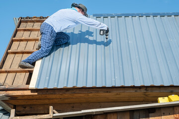 a rural male builder repairs the roof of a house, screws metal galvanized sheets of roofing iron with an electric cordless screwdriver