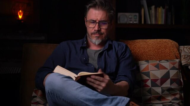 Happy man sitting on couch at home in cosy room reading book. Relaxing, leisure, education. Portrait of mature age, middle age, mid adult man, bearded, glasses, smiling, authentic look.