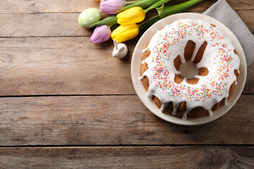 Glazed Easter cake with sprinkles, decorative bunny, painted egg and tulips on wooden table, flat lay. Space for text