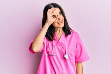 Middle age brunette woman wearing doctor uniform and stethoscope doing ok gesture with hand smiling, eye looking through fingers with happy face.