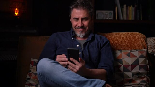 Man sitting on couch at home using phone. Businessman working in home office. Social media, newsfeed, chat. Mature age, middle age, mid adult man, bearded, authentic look.