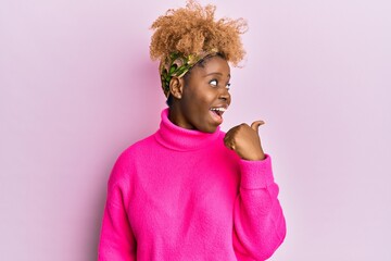 Young african woman with afro hair wearing casual winter sweater pointing thumb up to the side...