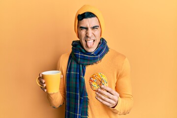 Handsome hispanic man eating doughnut and drinking coffee sticking tongue out happy with funny...