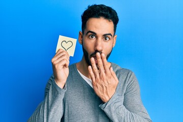 Young hispanic man holding heart reminder covering mouth with hand, shocked and afraid for mistake. surprised expression