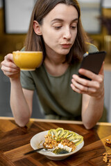 Woman in casual green t-shirt having a breakfast and chatting on smart phone during a morning time on the kitchen at home. Holding a cup of coffee an her hand.