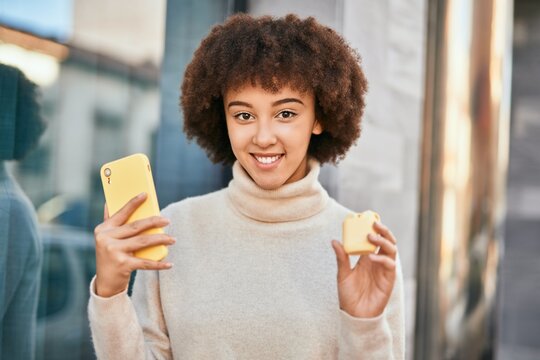 Young hispanic girl smiling happy using smartphone and holding earphones at the city.