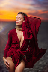 Beautiful blonde woman is posing in dress on desert with naked sexy legs. Red long silk elegant satin dress, slit up leg skirt, fluttering fly in wind motion. Summer photo. Fashion model, glamorous