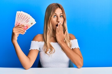 Beautiful blonde young woman holding 20 israel shekels banknotes covering mouth with hand, shocked...