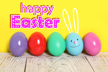 Happy Easter. Blue egg with drawn face and ears as bunny among others on white wooden table against yellow background