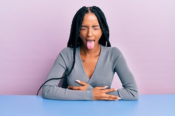 Beautiful hispanic woman wearing casual clothes sitting on the table sticking tongue out happy with...