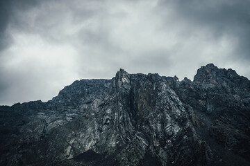 Dark atmospheric landscape with rocky mountain wall under gray cloudy sky. Rocky pinnacle in lead gray cloudy sky. Dark mountain peak in overcast weather. Gloomy minimalist scenery with high mountains