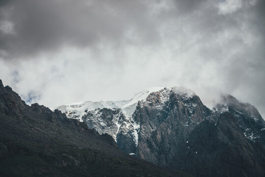 Dark atmospheric surreal landscape with dark rocky mountain top in low clouds in gray cloudy sky. Gray low cloud on high pinnacle. High black rock with snow in low clouds. Surrealist gloomy mountains.