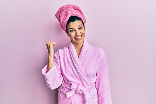 Young hispanic woman wearing shower towel cap and bathrobe smiling with happy face looking and pointing to the side with thumb up.