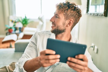 Handsome caucasian man smiling happy sitting on the sofa at home using touchpad device