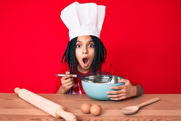 Young african american girl child with braids cooking using baker whisk scared and amazed with open...