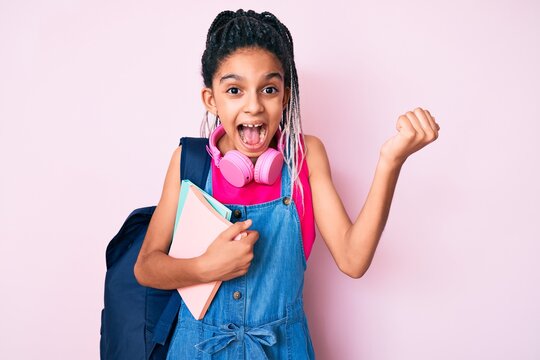Young african american girl child with braids holding student backpack and books screaming proud, celebrating victory and success very excited with raised arms