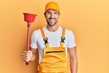 Young handsome man wearing plumber uniform holding toilet plunger looking positive and happy standing and smiling with a confident smile showing teeth