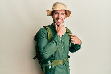 Handsome man with beard wearing explorer hat and backpack looking confident at the camera smiling with crossed arms and hand raised on chin. thinking positive.
