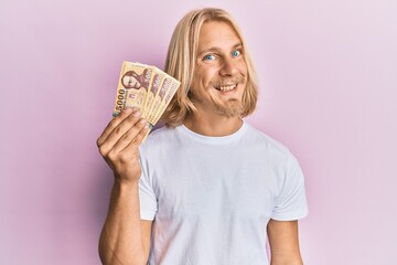 Caucasian young man with long hair holding 5000 hungarian forint banknotes looking positive and happy standing and smiling with a confident smile showing teeth