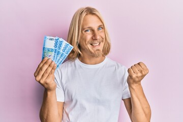 Caucasian young man with long hair holding 1000 hungarian forint banknotes screaming proud, celebrating victory and success very excited with raised arm