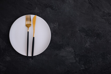 Empty plate with fork and knife on dark stone background. Gold and black tableware with white plate
