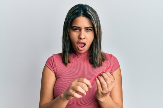Young latin woman using file nail in shock face, looking skeptical and sarcastic, surprised with open mouth