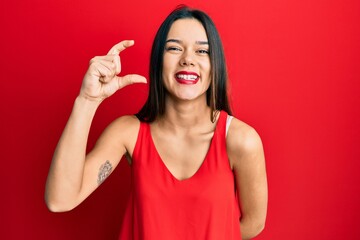 Young hispanic girl wearing casual style with sleeveless shirt smiling and confident gesturing with hand doing small size sign with fingers looking and the camera. measure concept.