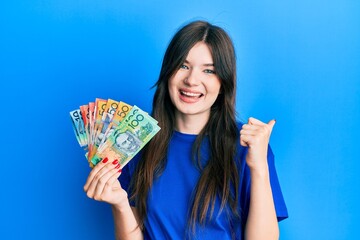 Young beautiful caucasian girl holding australian dollars pointing thumb up to the side smiling happy with open mouth