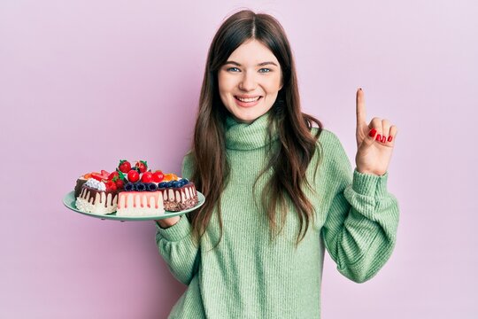 Young beautiful caucasian girl holding cake slices surprised with an idea or question pointing finger with happy face, number one