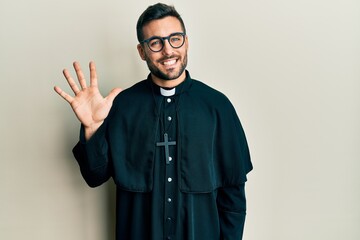 Young hispanic man wearing priest uniform standing over white background showing and pointing up with fingers number five while smiling confident and happy.