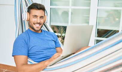 Young hispanic man relaxed working using laptop and headphones lying on the hammock at terrace.