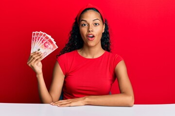 Young african american girl holding israel shekels sitting on the table scared and amazed with open...