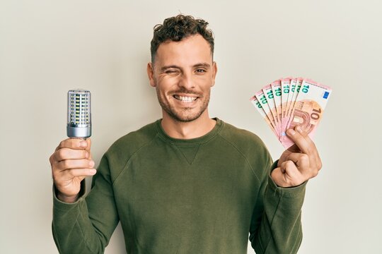 Young hispanic man holding led bulb and euros banknotes winking looking at the camera with sexy expression, cheerful and happy face.