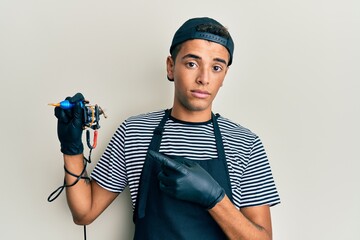 Young handsome african american man tattoo artist wearing professional uniform and gloves holding tattooer machine pointing with hand finger to the side showing advertisement, serious and calm face