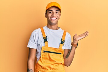 Young handsome african american man wearing handyman uniform over yellow background smiling cheerful presenting and pointing with palm of hand looking at the camera.