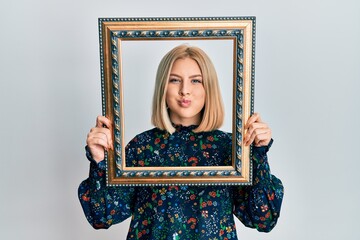 Fototapeta premium Young blonde woman holding empty frame puffing cheeks with funny face. mouth inflated with air, catching air.