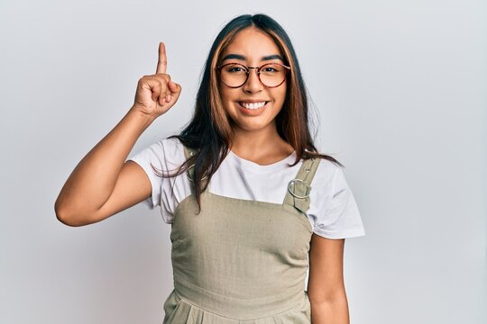 Young latin woman wearing casual clothes and glasses pointing finger up with successful idea. exited and happy. number one.