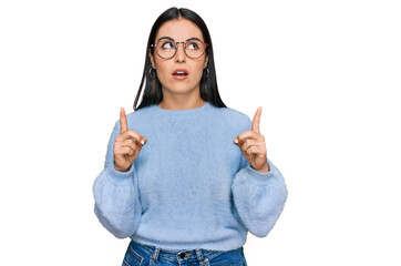 Young hispanic woman wearing casual clothes and glasses amazed and surprised looking up and pointing with fingers and raised arms.