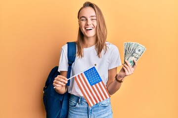 Beautiful blonde woman exchange student holding usa flag and dollars banknotes smiling and laughing...