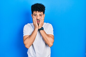 Young arab man with beard wearing casual white t shirt afraid and shocked, surprise and amazed expression with hands on face