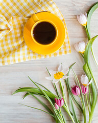 Morning coffee in bright yellow cup, napkin. Fresh garden pink and white tulips, spring flowers on white wooden background, top view from above