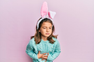 Little beautiful girl wearing cute easter bunny ears with hand on stomach because indigestion, painful illness feeling unwell. ache concept.