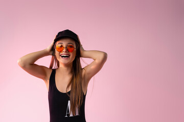 Portrait of a young tanned girl in a black swimsuit and sunglasses smiling happily. Neon pink background with side ad space.