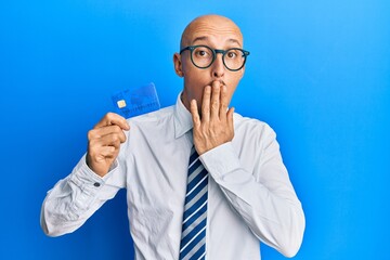 Bald man with beard holding credit card covering mouth with hand, shocked and afraid for mistake....