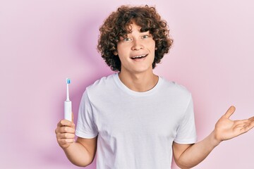 Handsome young man holding electric toothbrush celebrating achievement with happy smile and winner expression with raised hand