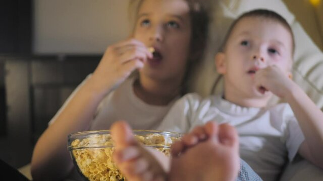 Happy family, brother and sister watching tv with popcorn bowl at home. Cute children eating popcorn while watching TV at home. Smiling kids watching funny cartoon together evening entertainment.