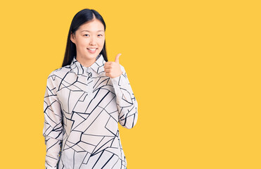 Young beautiful chinese woman wearing casual shirt doing happy thumbs up gesture with hand. approving expression looking at the camera showing success.
