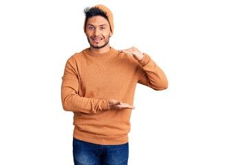 Handsome latin american young man wearing winter sweater and wool hat gesturing with hands showing big and large size sign, measure symbol. smiling looking at the camera. measuring concept.