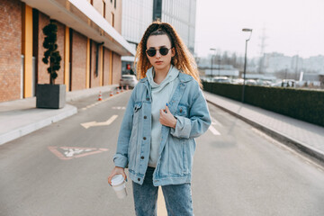 Young woman with a reusable cup of coffee, wearing denim jacket, outdoors, on city streets.